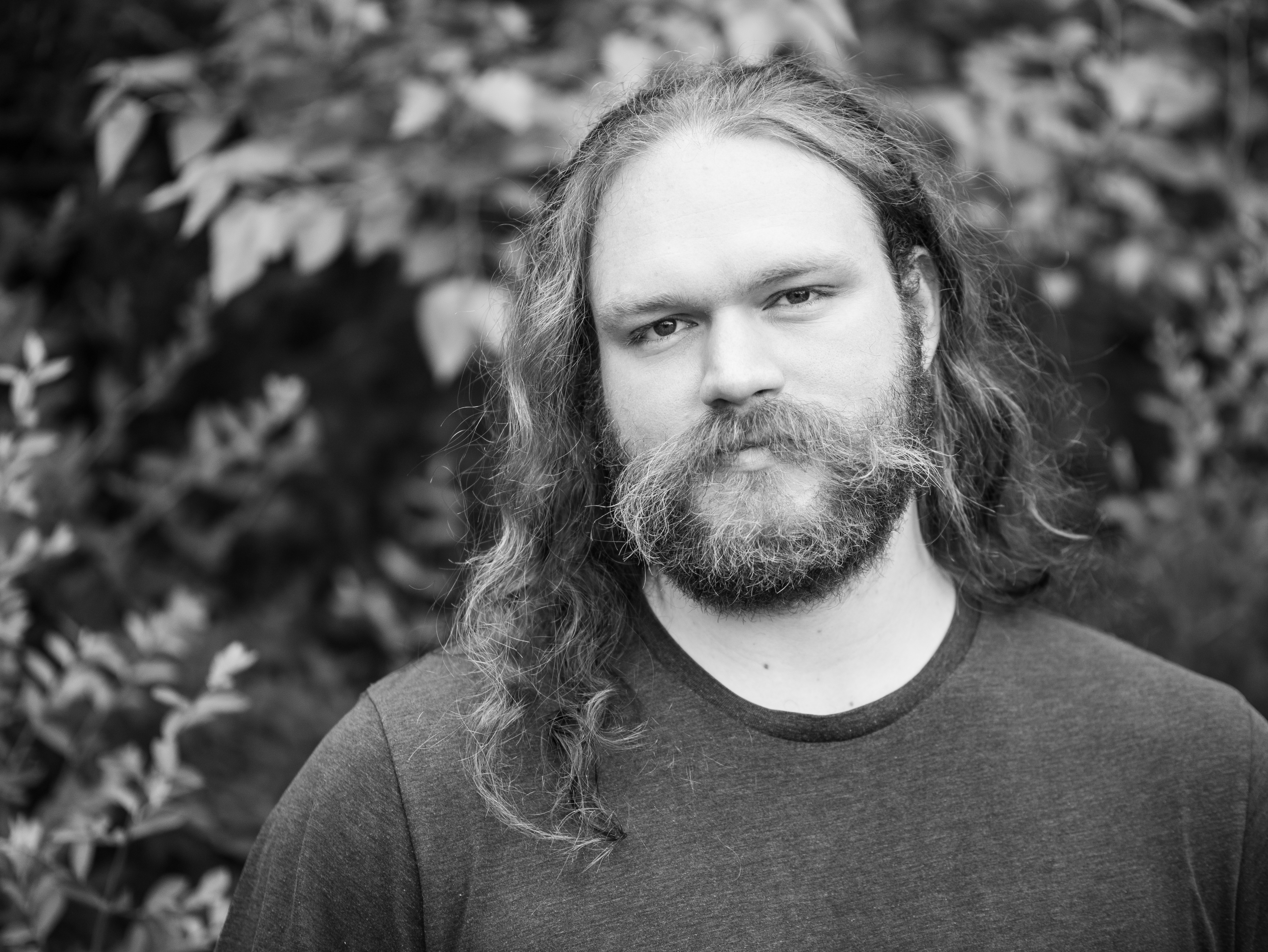 A black and white photograph of Doran. He has long blond hair, a beard, and a mustache. He is standing in front of a leafy tree wearing a t-shirt.